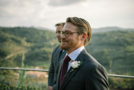 A man in a gray suit, red tie, and glasses with a white flower boutonnière smiles outdoors, with blurred greenery and mountains in the background. Another person in a suit stands behind him. by Justin Salem Meyer