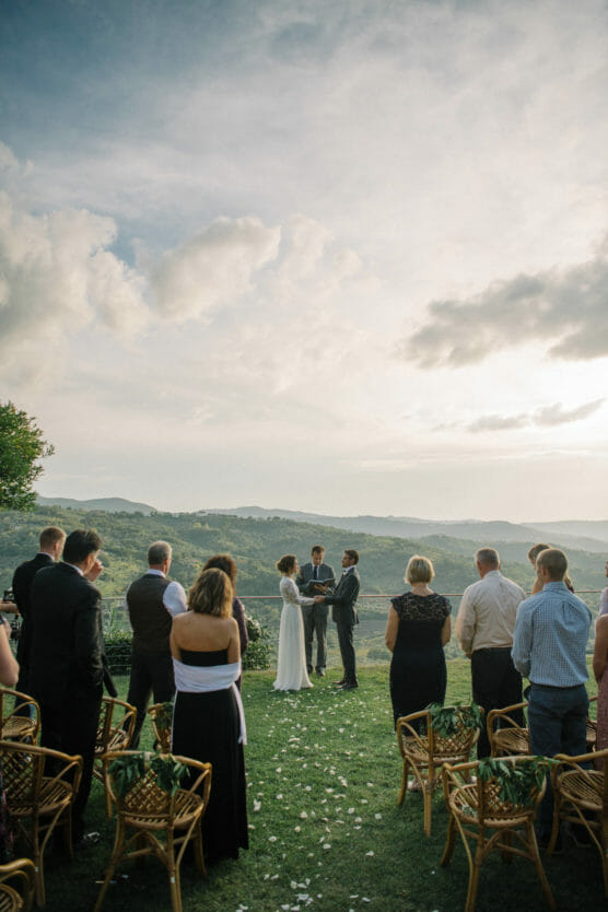 A couple stand facing each other during an outdoor wedding ceremony, with guests seated and standing on either side. The scene overlooks scenic green hills under a partly cloudy sky. by Justin Salem Meyer