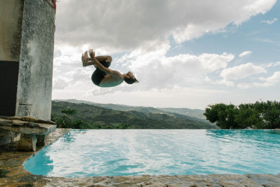 A person in mid-air performs a backflip off the edge of a stone pool, with hills and a cloudy sky in the background. by Justin Salem Meyer