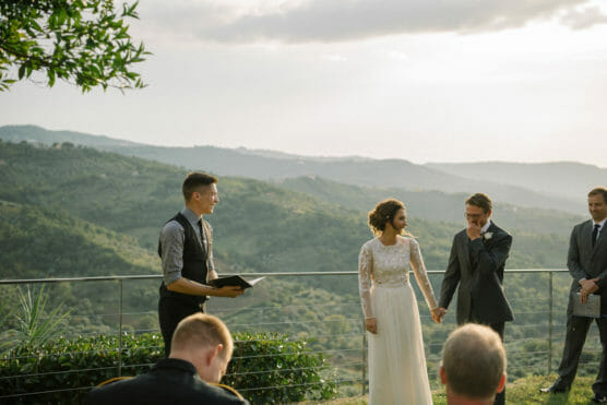 A bride and groom hold hands during an outdoor wedding ceremony on a scenic hillside, with mountains in the background. An officiant stands beside them, and a few guests watch in the foreground. by Justin Salem Meyer