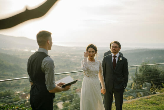 A bride in a white dress holds hands with a groom in a suit as they stand outdoors with an officiant, overlooking a scenic landscape at sunset. The bride is smiling and making a gesture with her hand. by Justin Salem Meyer