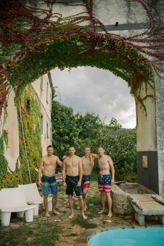 Four men in swim trunks stand smiling near a pool under an ivy-covered stone archway. The setting is outdoors beside an old building with greenery and overcast skies in the background. by Justin Salem Meyer