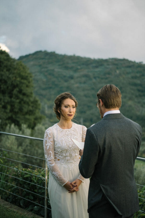 A bride in a white lace dress stands outdoors, listening to a groom in a suit reading vows, with green hills and cloudy sky in the background. by Justin Salem Meyer