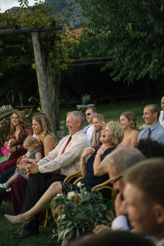 A group of people sit outdoors, laughing and smiling during a ceremony. Adults and children are seated on wooden chairs, surrounded by greenery and trees, with mountains visible in the background. by Justin Salem Meyer