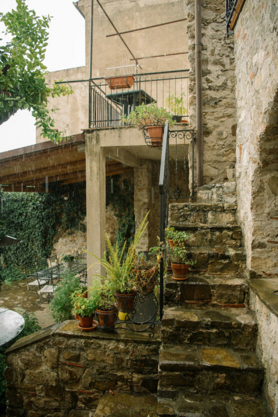 A stone staircase with potted plants leads to a terrace in a rustic courtyard. Ivy climbs the walls, and rain falls gently, creating a serene, lush atmosphere. by Justin Salem Meyer