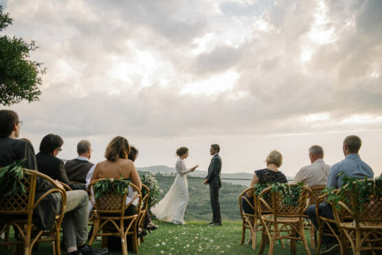 A bride and groom stand facing each other outdoors on a grassy hilltop, surrounded by guests seated in decorated chairs, with a scenic landscape and cloudy sky in the background. by Justin Salem Meyer
