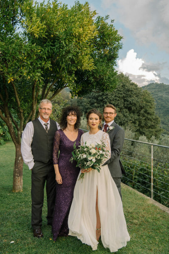 Four people pose outdoors at a wedding: a bride in a white dress holds a bouquet, standing beside a groom in a suit, and two older adults in formal attire. Lush greenery and hills fill the background. by Justin Salem Meyer
