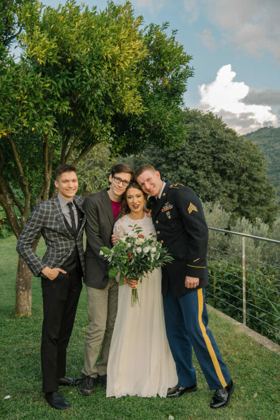 Four people stand together outdoors in front of trees and hills. The woman in the center wears a white dress and holds a bouquet. Three men stand close around her, one in a military uniform, all smiling happily. by Justin Salem Meyer