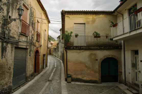 A narrow cobblestone street winds between old, weathered buildings with shuttered windows, potted plants on balconies, and soft, muted yellow and brown walls in a quiet, historic European town. by Justin Salem Meyer