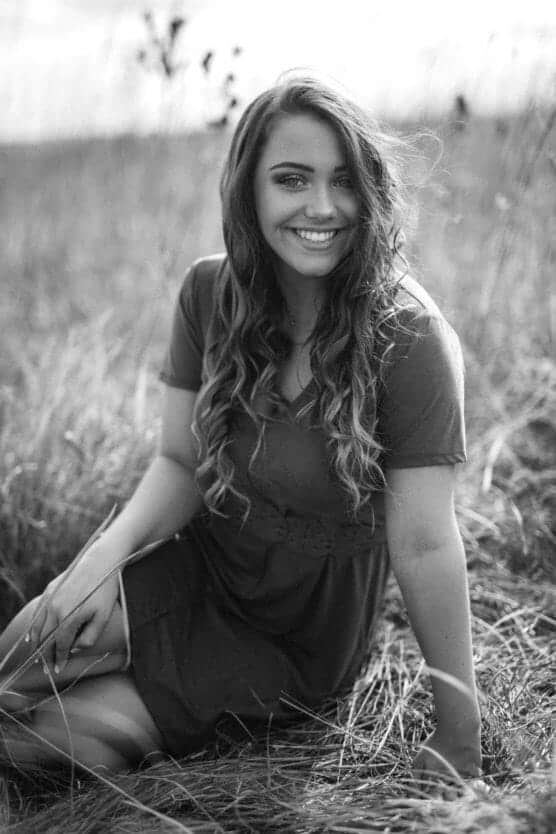 A young woman with long wavy hair sits smiling in a grassy field, wearing a short-sleeved dress. The black and white photo captures a relaxed and joyful mood in natural light. by Justin Salem Meyer