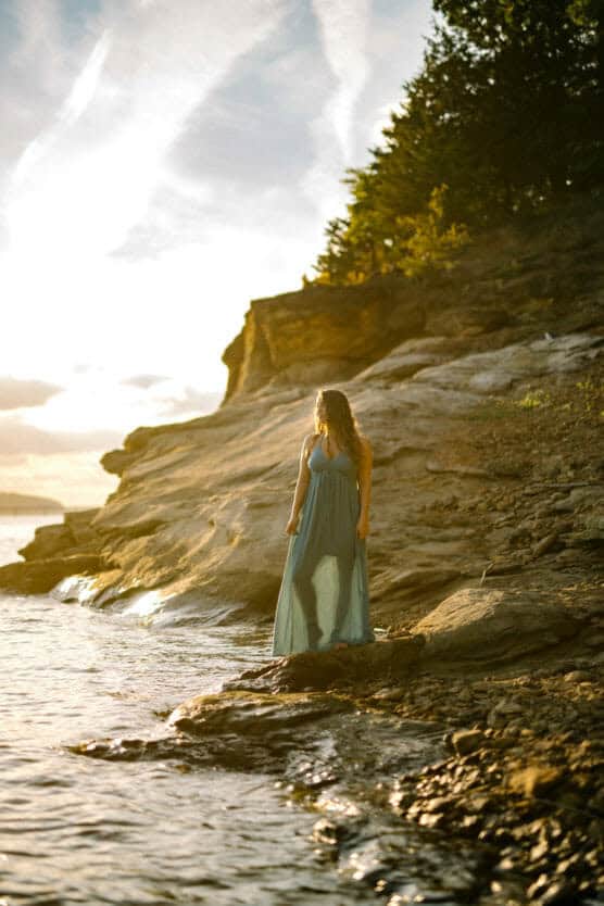 A woman in a flowing blue dress stands on rocky terrain by the waters edge at sunset, looking into the distance with sunlight illuminating the scene. by Justin Salem Meyer