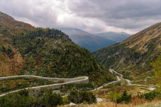 A winding mountain road curves through a lush, green valley surrounded by forested hills and distant peaks under a cloudy, overcast sky. by Justin Salem Meyer