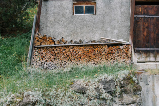 A stack of firewood is neatly arranged against the concrete wall of a rustic building with a small window and wooden door, surrounded by green grass and vegetation. by Justin Salem Meyer