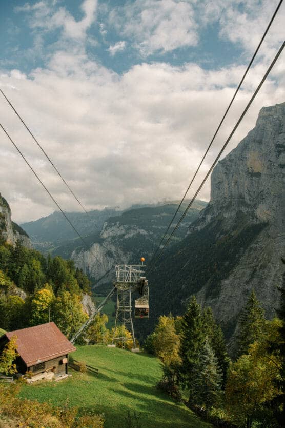 A cable car travels above a green alpine valley with steep rocky cliffs, dense trees, and a rustic wooden cabin. The sky is partly cloudy with mountains in the background. by Justin Salem Meyer