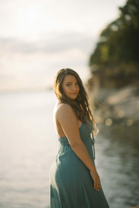 A woman with long wavy hair wearing a sleeveless teal dress stands by the water’s edge, looking back over her shoulder with a soft smile. The background is bright and blurred, with trees and shoreline visible. by Justin Salem Meyer