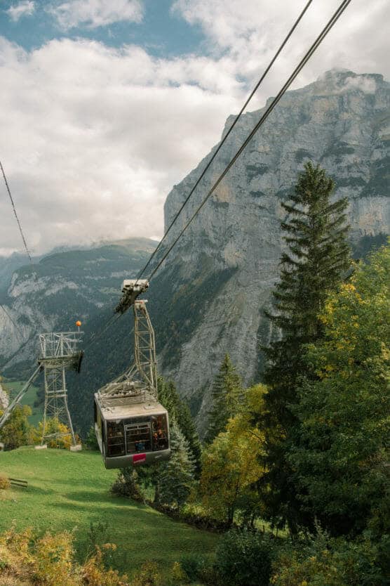A cable car ascends a mountain, passing over green grass and trees, with a backdrop of steep cliffs and cloudy skies in a scenic alpine landscape. by Justin Salem Meyer