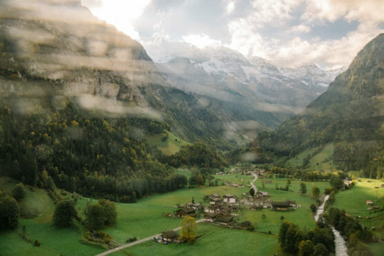 A picturesque valley with a small village, green fields, scattered trees, and a winding river, surrounded by tall mountains with snowy peaks under a partly cloudy sky. by Justin Salem Meyer