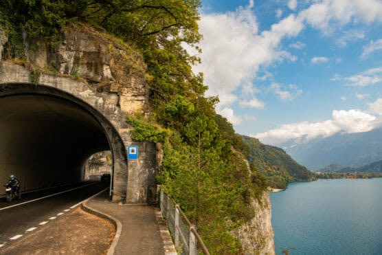 A tunnel carved into a rocky cliff beside a lake, with a motorcyclist exiting the tunnel, a walking path, green trees, and mountains in the background under a partly cloudy sky. by Justin Salem Meyer