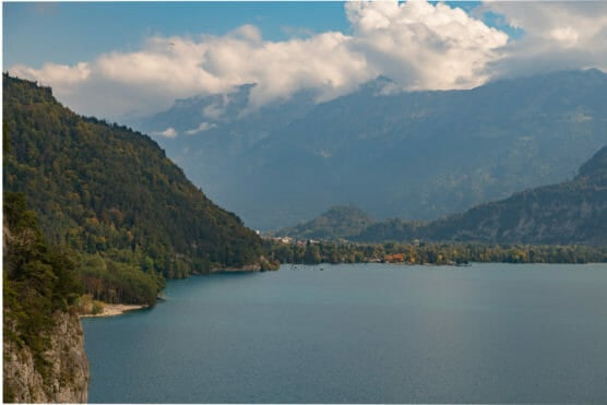 A serene lake bordered by forested hills and mountains under a partly cloudy sky, with a few distant boats and a small area of colorful trees along the shoreline. by Justin Salem Meyer