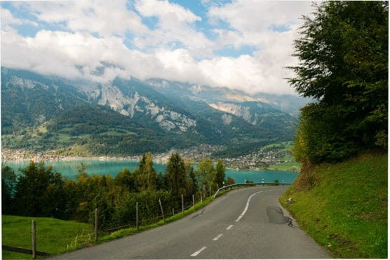 A winding road descends toward a turquoise lake, bordered by lush green hills, with a village and forested mountains in the background under a partly cloudy sky. by Justin Salem Meyer