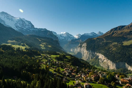 A scenic mountain valley with a village nestled among green hills, surrounded by forested slopes and snow-capped peaks under a clear blue sky. by Justin Salem Meyer