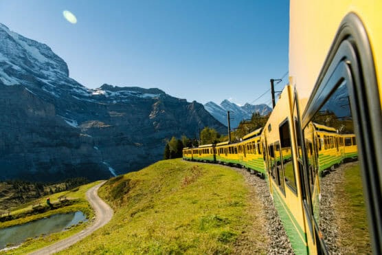 A yellow train travels through green mountain scenery, with snow-capped peaks and a clear blue sky in the background. The trains reflection is visible in its windows as it curves along the track. by Justin Salem Meyer