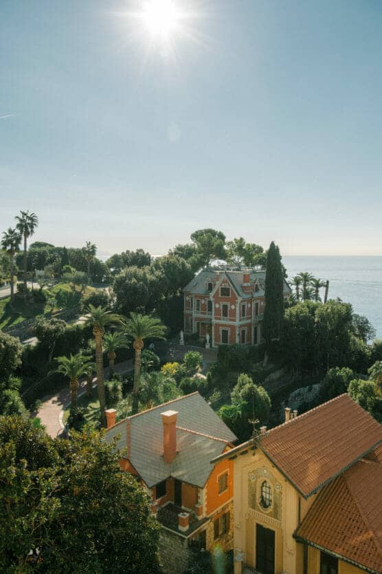 A sunlit coastal scene features a large red villa surrounded by lush greenery and palm trees, with the sea visible in the background and several smaller buildings with terracotta roofs in the foreground. by Justin Salem Meyer