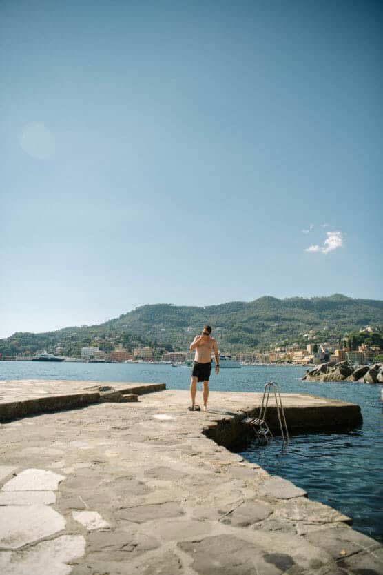 A man in swim trunks stands on a stone pier by the water, with mountains and a coastal town in the background under a clear blue sky. by Justin Salem Meyer
