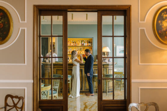 A couple dressed formally stands facing each other by a bar in an elegant, warmly lit room, seen through wooden-framed glass doors. The bar is stocked with bottles, and the decor features classic paintings and refined furnishings. by Justin Salem Meyer