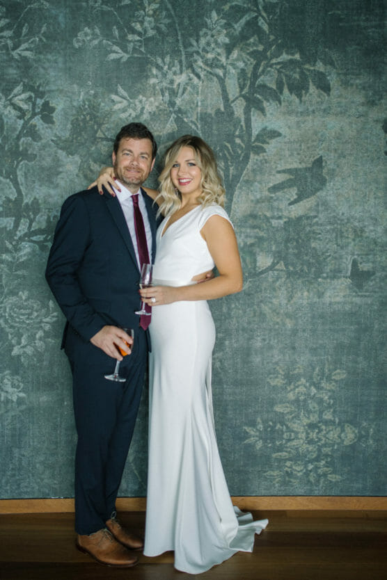 A man in a dark suit and a woman in a white gown stand together smiling, each holding a drink, in front of a green floral-patterned backdrop. by Justin Salem Meyer