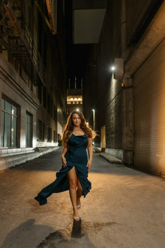 A woman in a dark, elegant dress walks confidently down a dimly lit urban alley at night, with her hair flowing and one leg revealed by a high slit in her dress. by Justin Salem Meyer