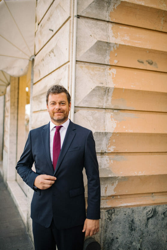 A man with short brown hair and a beard, wearing a navy blue suit, white shirt, and red tie, stands smiling near a textured, weathered wall on a city sidewalk. by Justin Salem Meyer