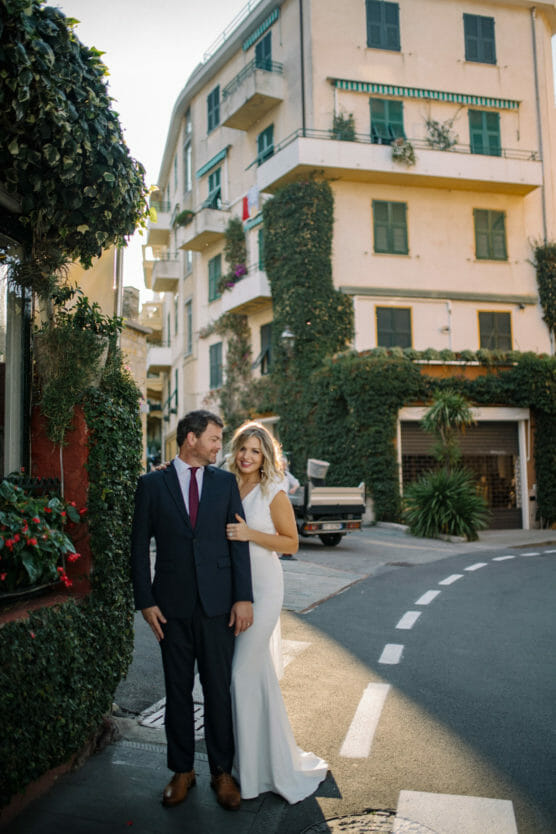 A smiling couple dressed in wedding attire stands on a sunlit street corner, with a beige, ivy-covered apartment building in the background. The bride wears a white dress and the groom wears a dark suit and tie. by Justin Salem Meyer