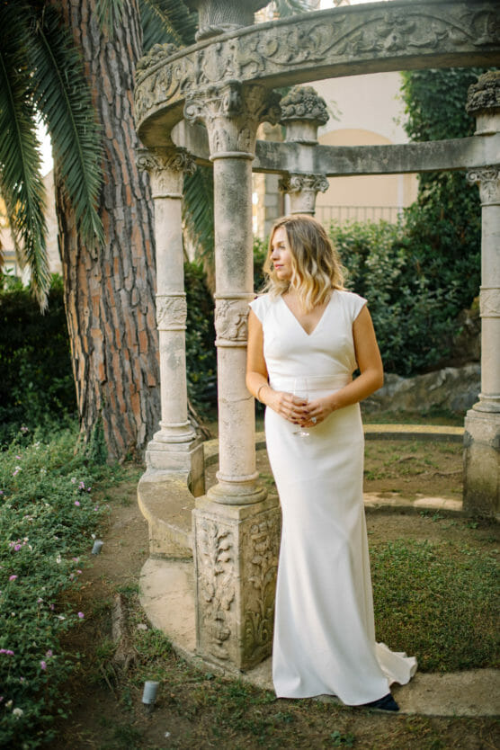 A woman in a white, floor-length dress stands by an ornate stone gazebo in a lush garden, gazing thoughtfully to the side, hands gently clasped in front of her. by Justin Salem Meyer