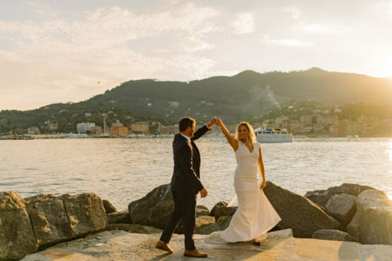 A couple, dressed in formal attire, dances joyfully on rocks by the water at sunset, with mountains and a town in the background. The woman wears a white dress, and the man wears a dark suit. A boat is visible on the water. by Justin Salem Meyer