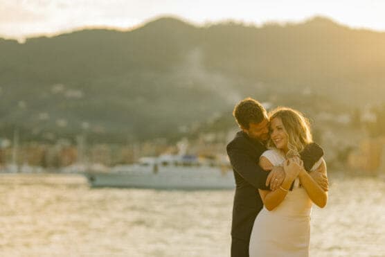 A man in a dark suit embraces a smiling woman in a light dress by the water at sunset, with a blurred yacht and mountains in the background. by Justin Salem Meyer