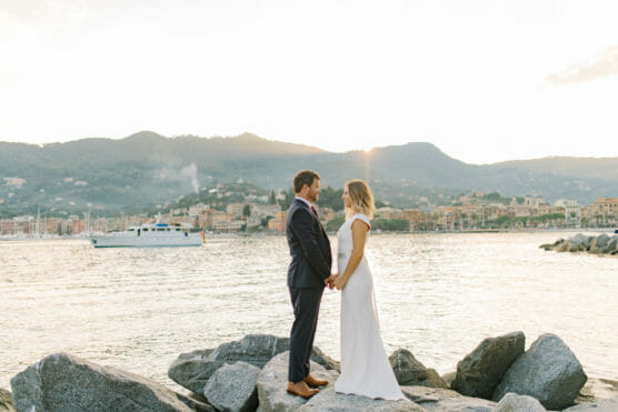 A couple in wedding attire stands barefoot on large rocks by the water, holding hands and facing each other, with a boat and a coastal town backed by mountains in the background at sunset. by Justin Salem Meyer