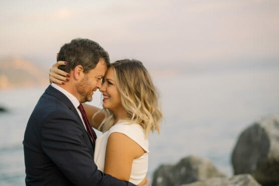 A smiling couple embraces closely by the water, with the woman holding the mans head and both looking happy. The background shows a calm, scenic shoreline at sunset. by Justin Salem Meyer