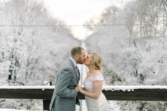 A bride and groom share a kiss outdoors on a snowy day, holding hands and standing on a wooden bridge surrounded by trees covered in snow. by Justin Salem Meyer