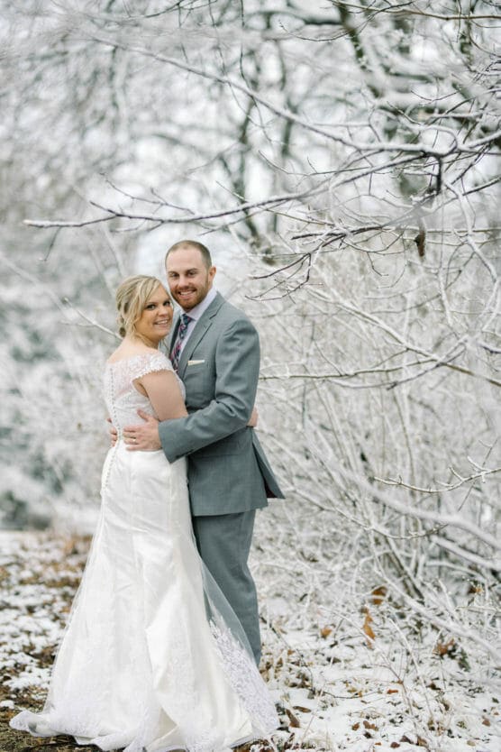 A bride in a white gown and a groom in a gray suit embrace and smile at the camera in a snowy, wintery outdoor setting with frost-covered trees and ground. by Justin Salem Meyer