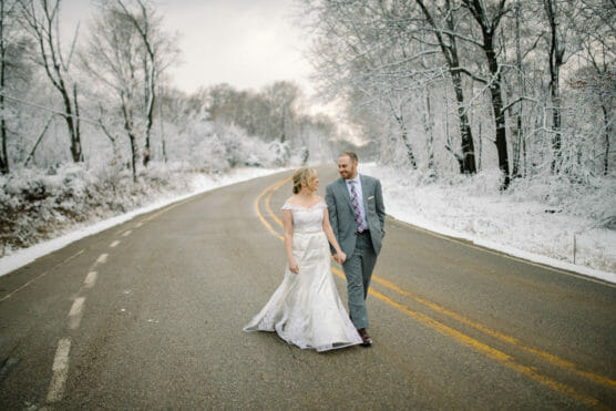 A bride in a white gown and a groom in a gray suit walk hand-in-hand on a winding road lined with snow-covered trees, creating a serene winter wedding scene. by Justin Salem Meyer