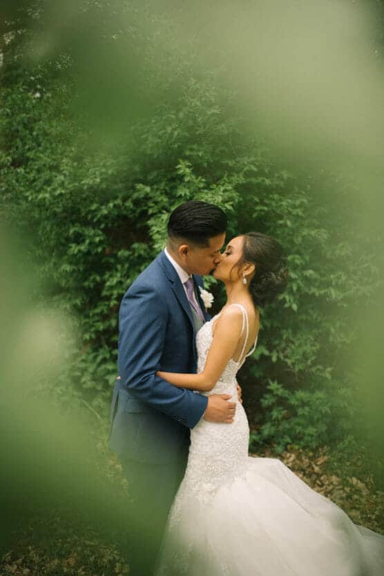 A bride and groom share a kiss outdoors, surrounded by lush green foliage. The bride wears a white lace gown, and the groom is in a blue suit. Blurry leaves in the foreground frame the romantic moment. by Justin Salem Meyer