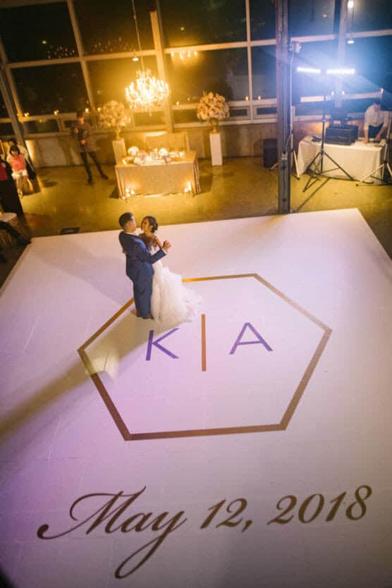 A bride and groom share their first dance on a white dance floor with a hexagon and the initials KIA. May 12, 2018 is written in script on the floor; a chandelier and decorated table are visible in the background. by Justin Salem Meyer