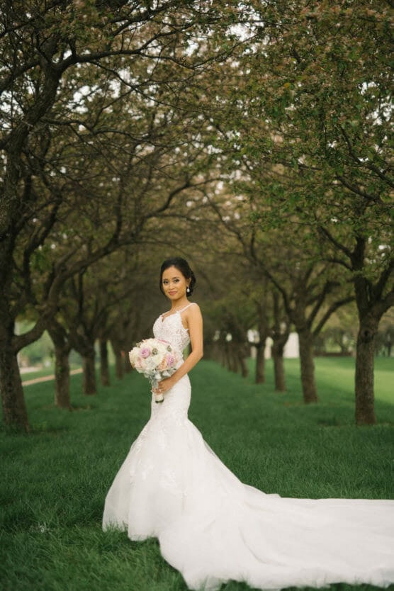 A bride in a white gown holds a bouquet of flowers while standing on green grass beneath a canopy of trees in bloom, creating a romantic outdoor wedding scene. by Justin Salem Meyer