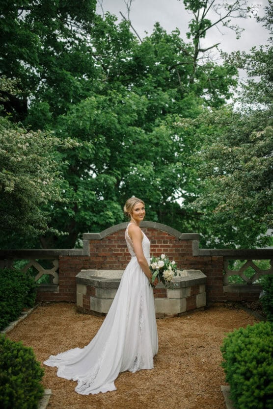 A bride in a white wedding dress stands outdoors on a gravel path, holding a bouquet of flowers. She faces away but looks over her shoulder, surrounded by green trees and bushes, with a brick wall and stone bench behind her. by Justin Salem Meyer