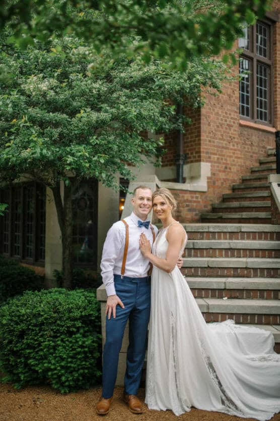 A bride in a white gown and a groom in a white shirt, blue pants, and suspenders stand smiling together outdoors by a brick building and greenery, with stairs in the background. by Justin Salem Meyer