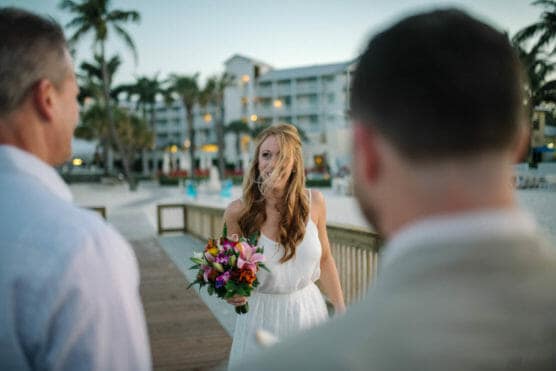 A woman in a white dress holding a colorful bouquet stands on a wooden walkway by the beach, wind blowing her hair across her face, with two men in dress shirts in the foreground. A resort and palm trees are in the background. by Justin Salem Meyer