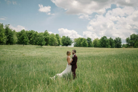 A bride and groom stand together in a grassy field under a partly cloudy sky, surrounded by green trees, sharing an intimate moment on their wedding day. by Justin Salem Meyer