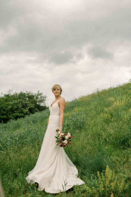 A woman in a white wedding dress stands in a grassy field under a cloudy sky, holding a bouquet of flowers and looking at the camera. by Justin Salem Meyer