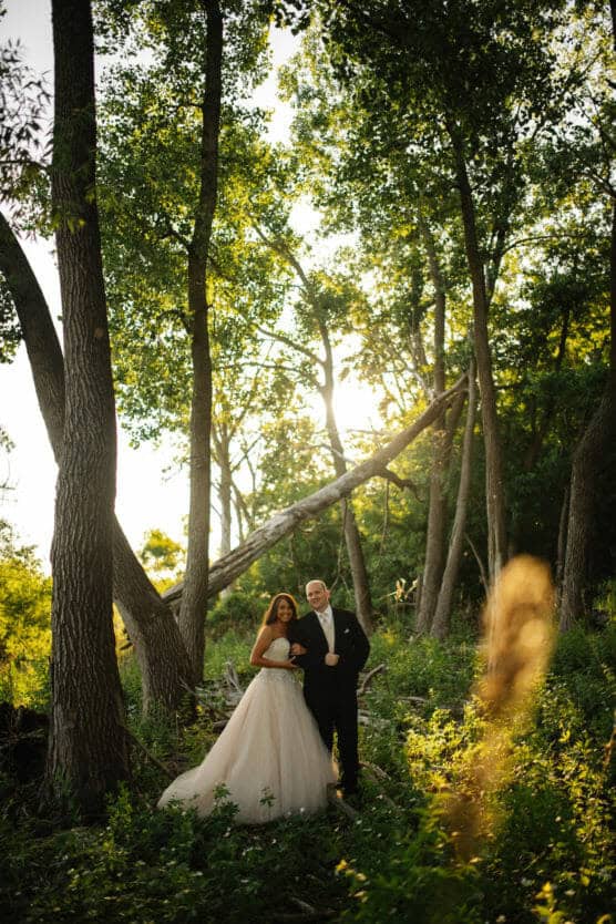 A bride and groom stand together in a sunlit forest, surrounded by tall trees and greenery. Sunlight filters through the leaves, casting a warm glow on the couple as they smile and pose in their wedding attire. by Justin Salem Meyer
